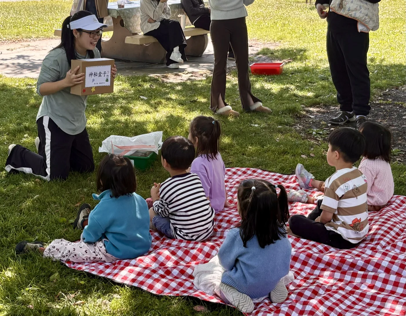 Ning presenting a mystery box activity to children seated on a picnic blanket
